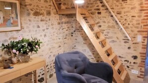 A rustic room with exposed wooden beams, a stone wall, a wooden ladder, a blue armchair, a wooden table, and a hanging light fixture. - Maison de Campagne (Moulins)