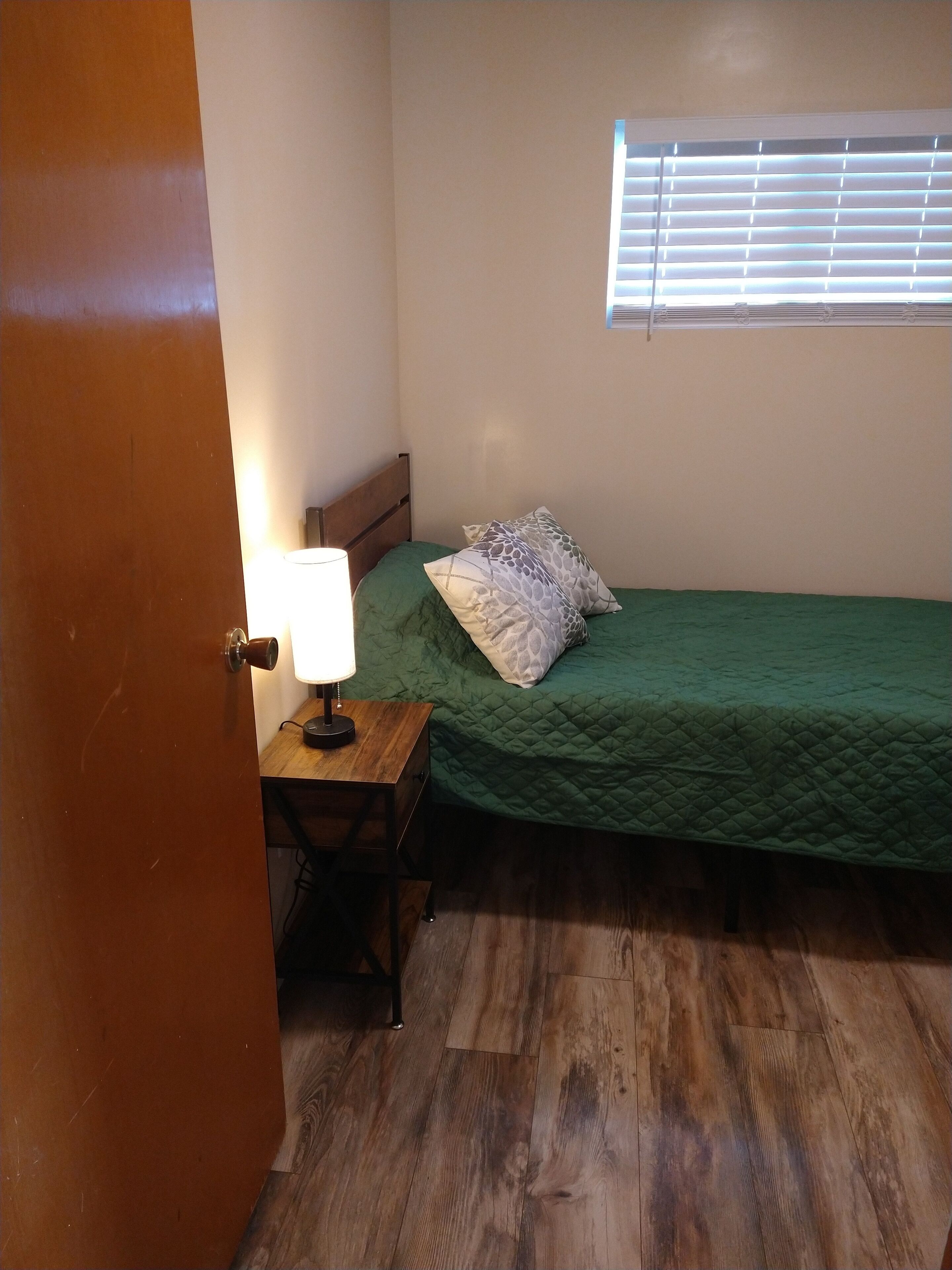Une petite chambre avec un lit simple, un plancher en bois, une table de chevet avec une lampe et une fenêtre avec des stores.
