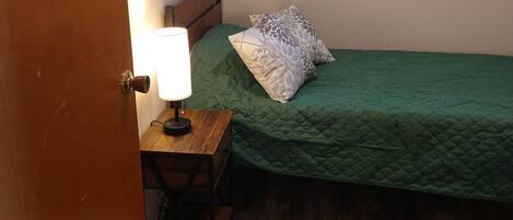 Une petite chambre avec un lit simple, un plancher en bois, une table de chevet avec une lampe et une fenêtre avec des stores.