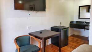 A compact room with a flat-screen TV mounted on the wall, a small table with a chair, a compact refrigerator, and a kitchenette with a sink and countertop.