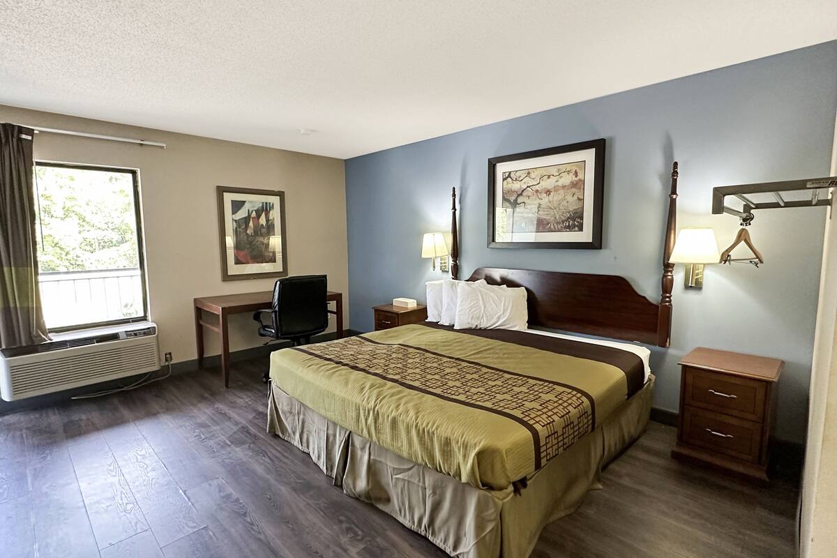 A hotel room with a bed, a desk, a chair, a nightstand, a lamp, and a window with curtains.