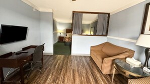 A hotel room with a wooden floor, a TV mounted on the wall, a desk with a chair, a striped sofa, a small table with a lamp, and a mirror with curtains.