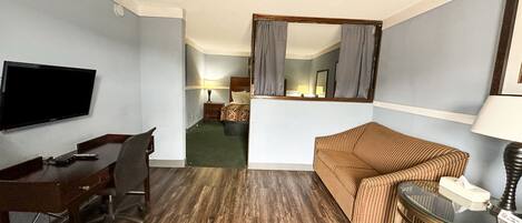 Quarto de hotel com piso de madeira, televisão montada na parede, escrivaninha com cadeira, sofá listrado, mesa pequena com abajur e espelho com cortinas.
