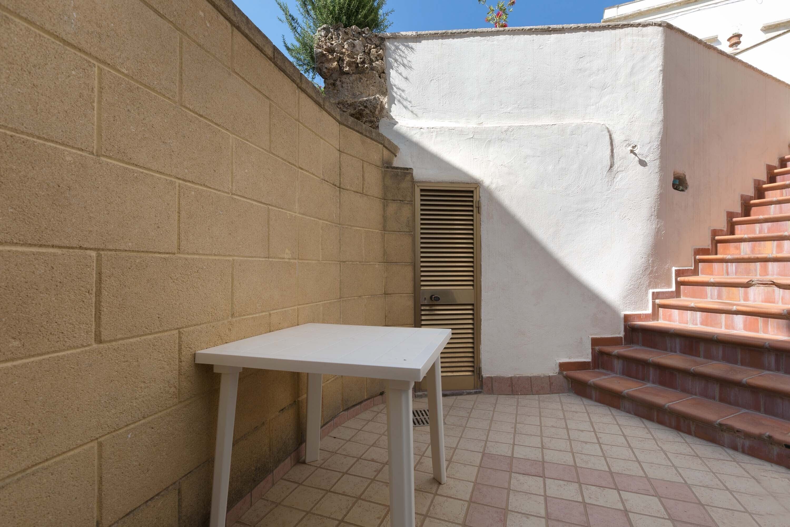 Un pequeño patio al aire libre con una mesa blanca, un muro de ladrillo y una escalera de baldosas rojas que conduce a una puerta.
