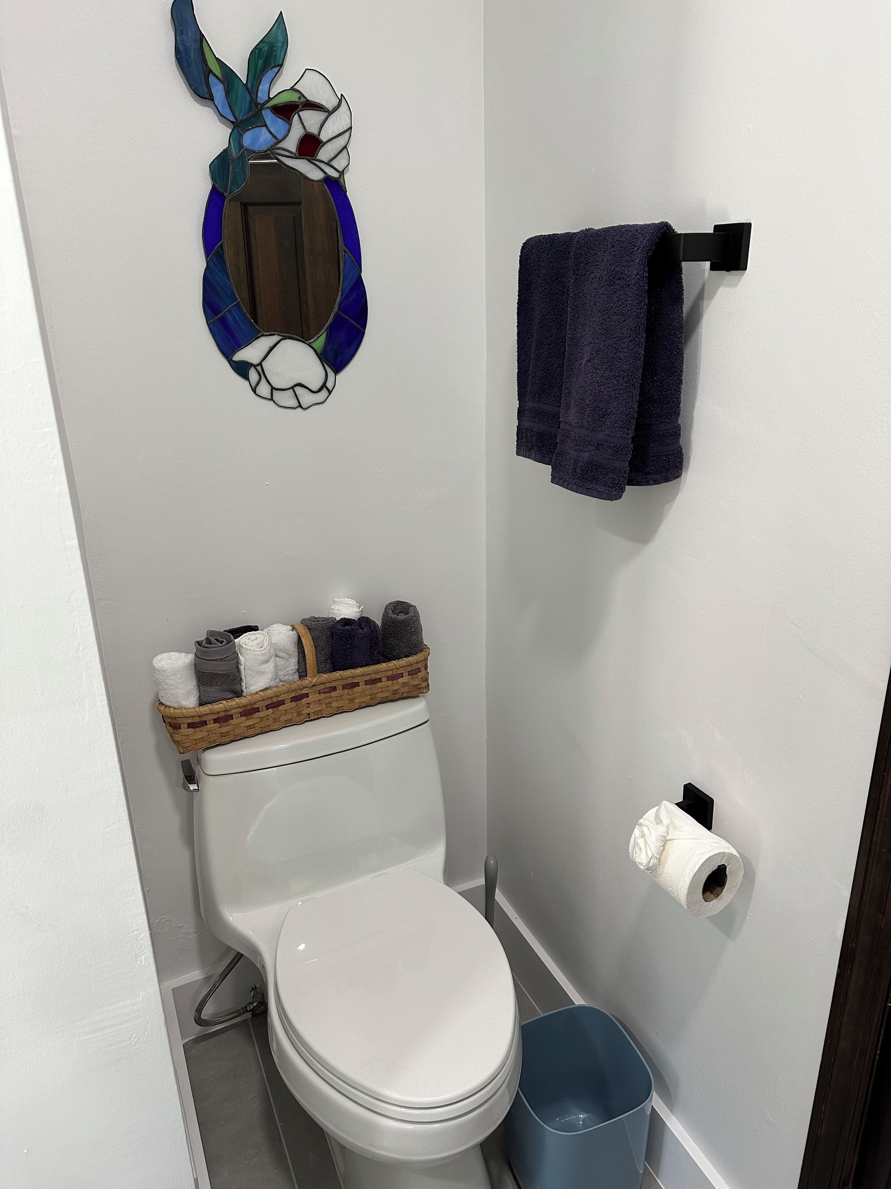 Un baño con inodoro, una cesta de toallas, dispensador de papel higiénico y un espejo decorativo.