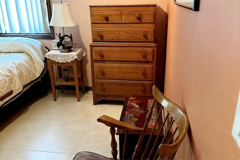 A bedroom with a bed, a wooden dresser, a small table with a lamp, and a chair.