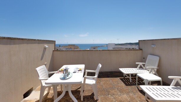 A rooftop terrace with a table set for two, white plastic chairs, and a view of the sea and buildings in the distance. - 3500 Panorama D`otranto by Barbarhouse (Otranto)
