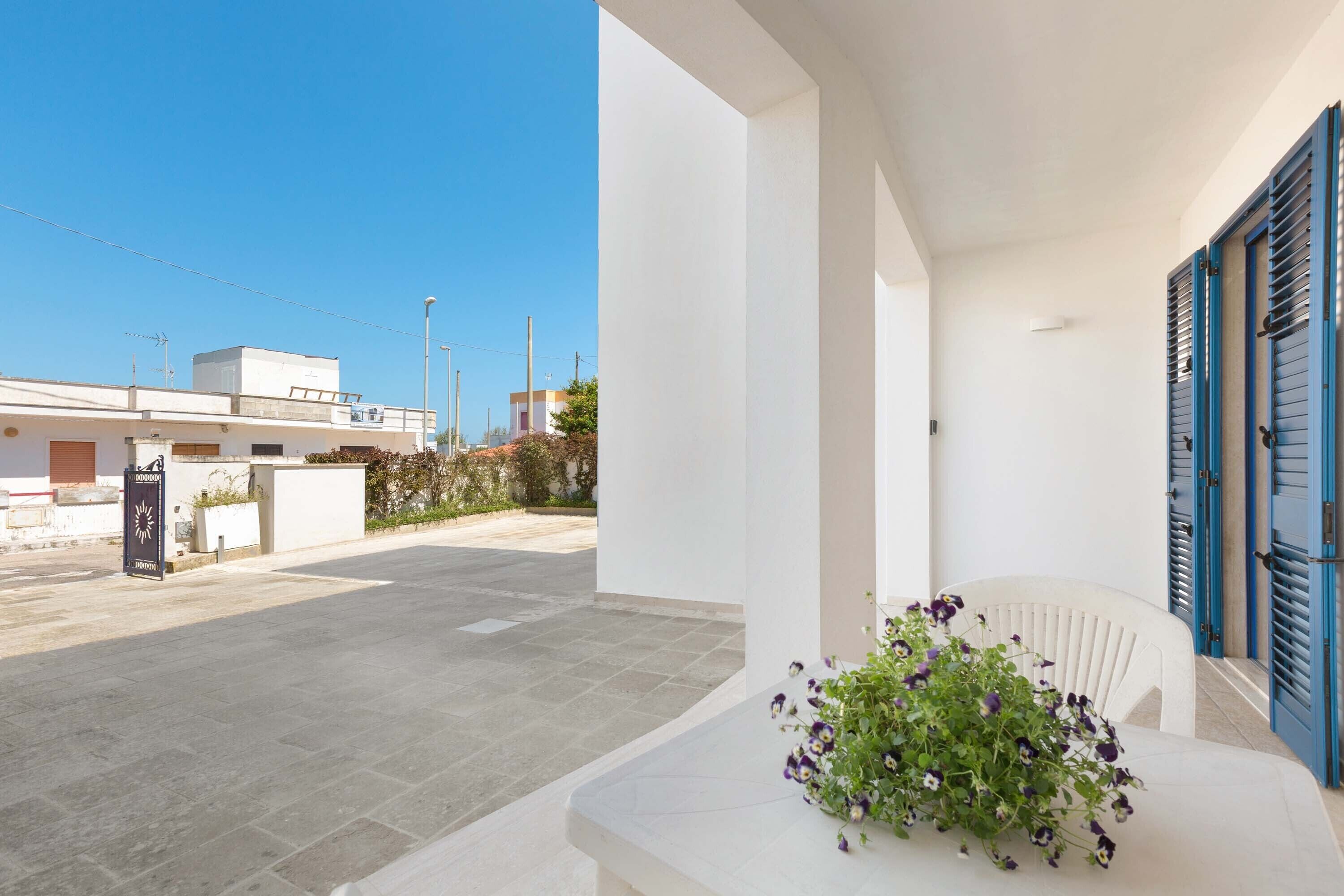 Un bâtiment moderne à la façade blanche et aux volets bleus, doté d’une cour pavée et agrémenté d’un arrangement floral posé sur un banc blanc.