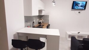 Un espacio habitable compacto con kitchenette, comedor con taburetes de bar, televisor de pantalla plana en la pared y un pequeño rincón de descanso con sofá estampado.
