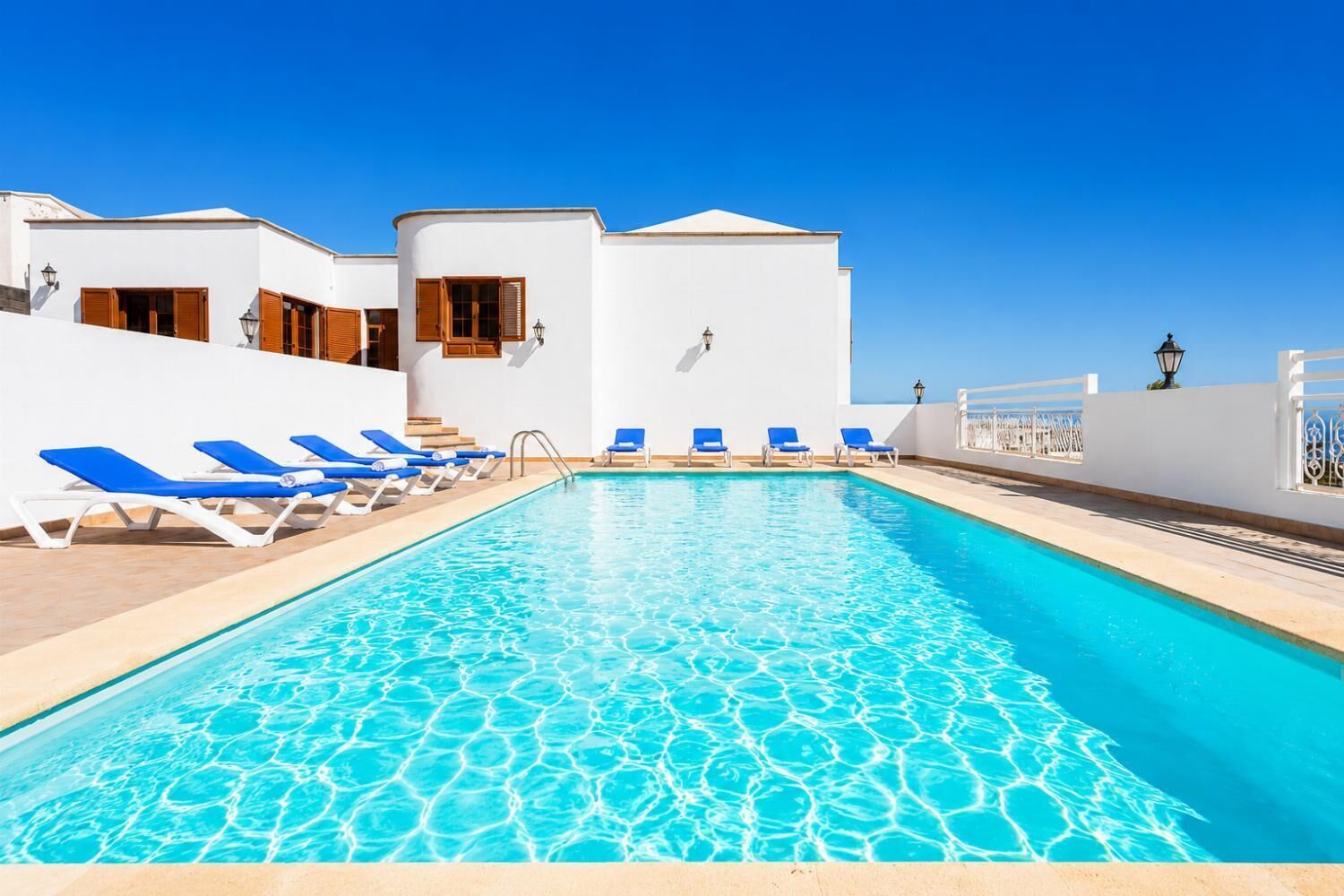 A pool with clear blue water, surrounded by white buildings with blue lounge chairs.