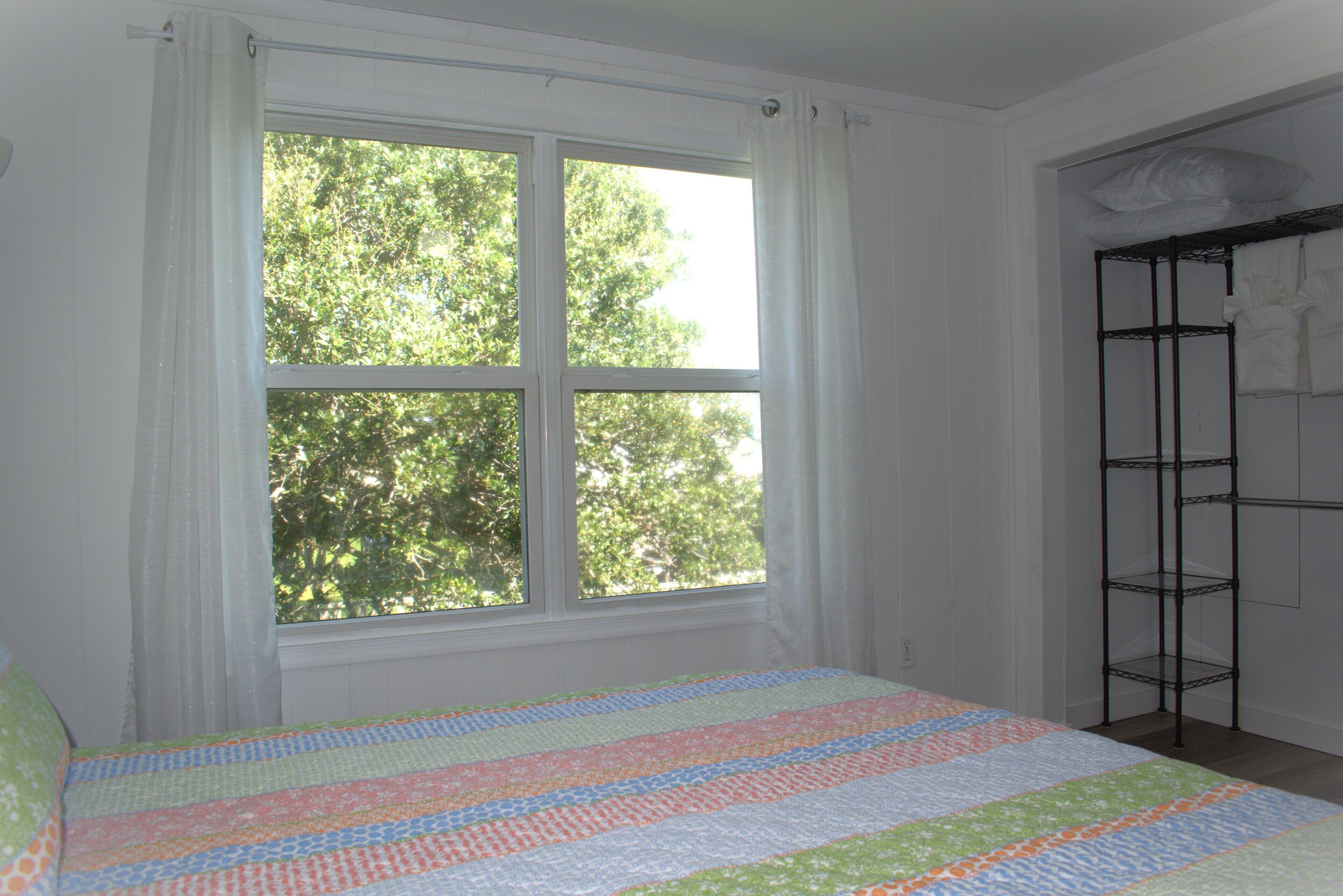 A bedroom with a large window, a bed with a colorful quilt, and a metal shelf unit against the wall.