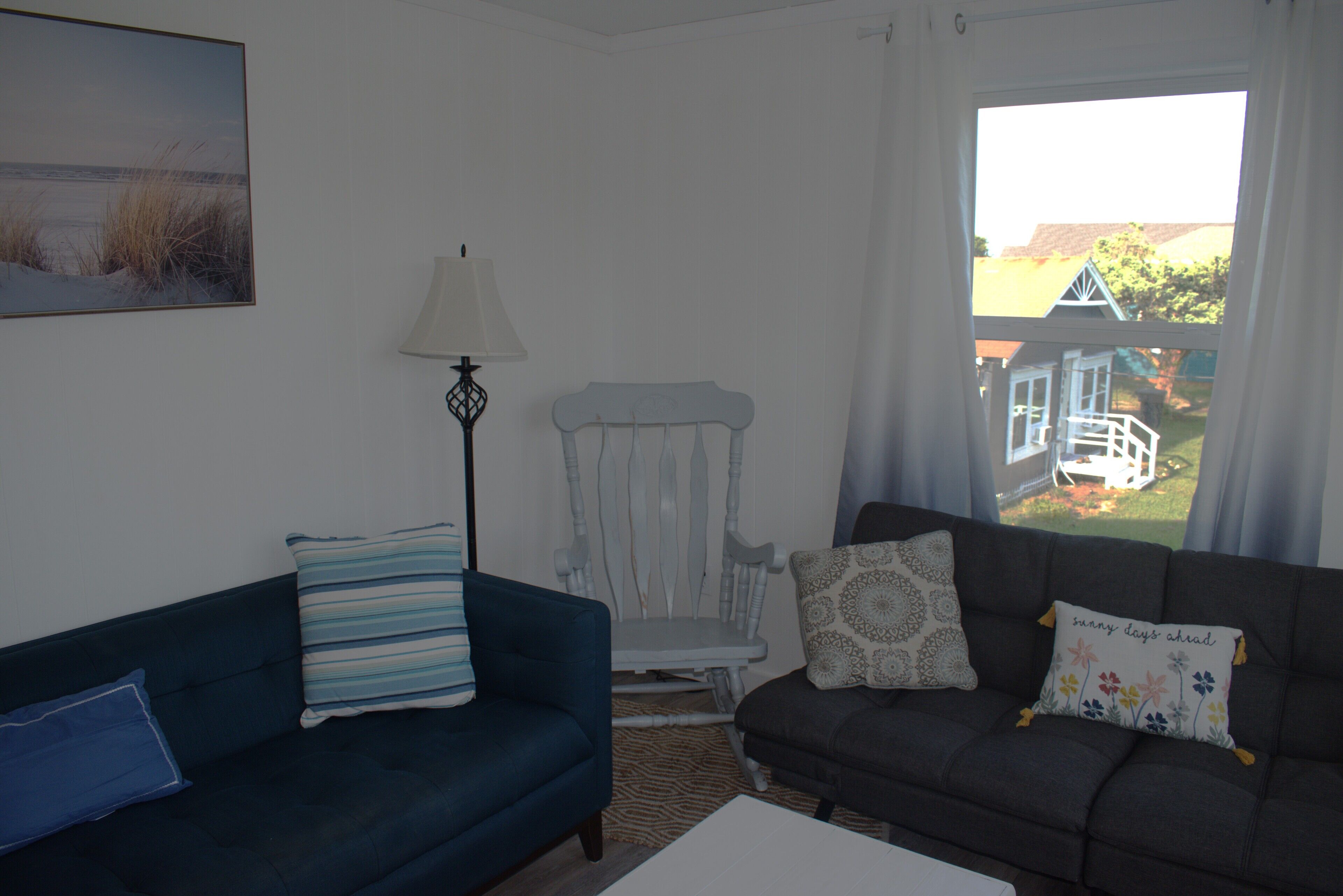 A living room with a blue sofa, a white chair, and a dark sofa with floral cushions. There is a lamp and a painting on the wall.