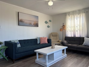 A living room with a dark blue sofa, a white coffee table, a beige armchair, and a grey sofa. There is a painting on the wall and a floor lamp. - Tranquil Tides Retreat in Frisco, Outer Banks, North Carolina! (Frisco)