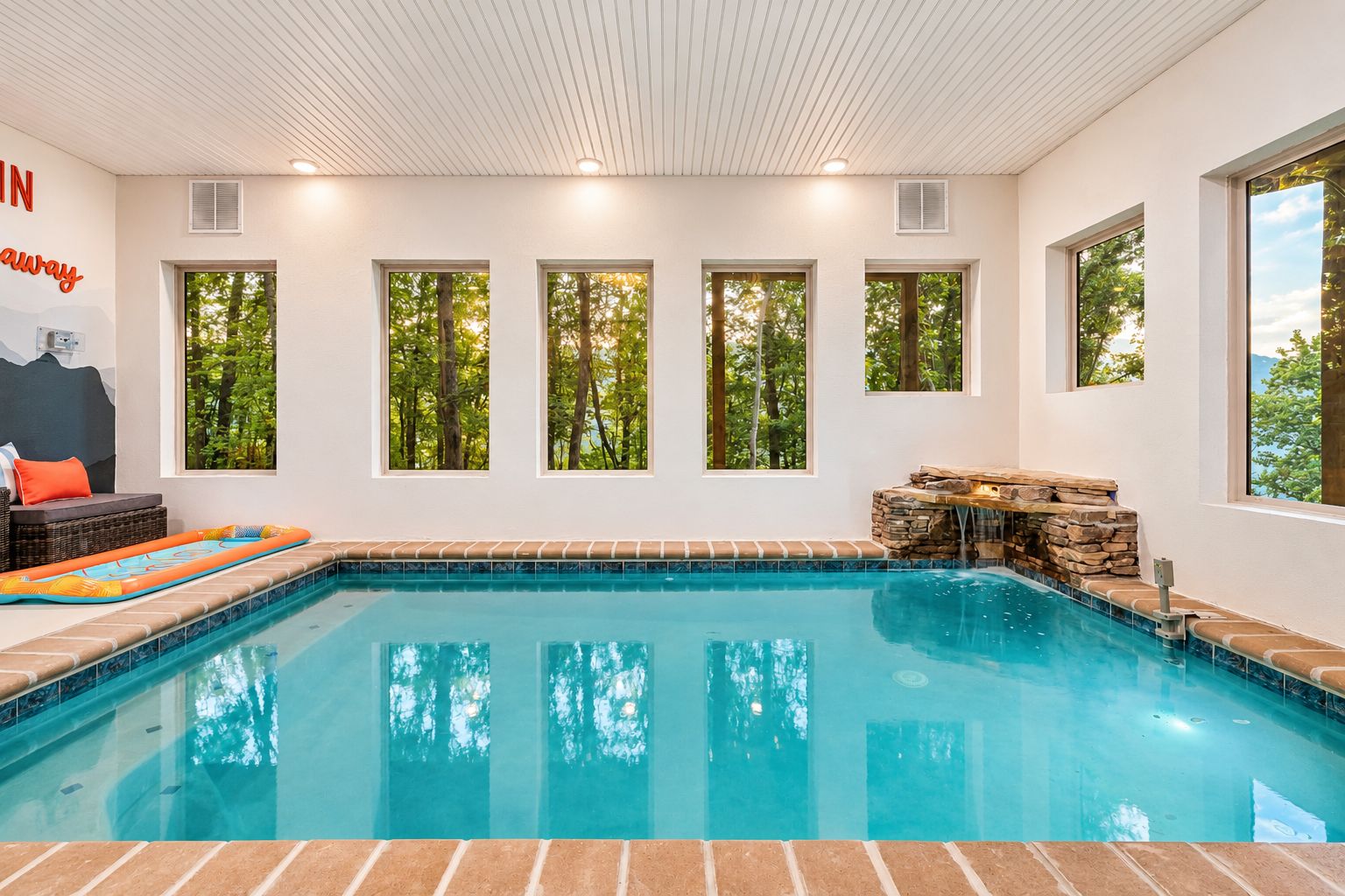 An indoor swimming pool with a brick border, multiple windows, and a stone waterfall feature.