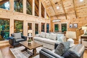 A spacious living area with a large sofa, a coffee table, and a fireplace. There are multiple windows providing natural light and views of the surrounding forest.