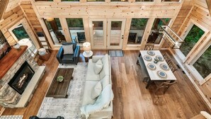 A spacious interior with a high ceiling, wooden beams, a fireplace, a sofa, a dining table, and large windows offering a view of the outdoors. - FreeDollywoodTix,HeatedPool,Luxurious,HotTub,Views (Sevierville)