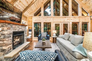 A spacious living area with a stone fireplace, wooden beams, and large windows offering views of the outdoors.