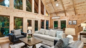 A spacious wooden cabin interior with a large dining area, a sofa set, and a view of the surrounding forest. - FreeDollywoodTix,HeatedPool,Luxurious,HotTub,Views (Sevierville)