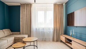 A living room with a sofa, two round tables, a TV on the wall, and sheer curtains.
