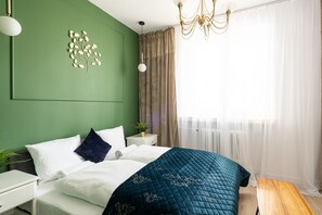 Une chambre à coucher bien rangée, avec un lit, des tables de chevet, un luminaire fixé au mur et une fenêtre ornée de rideaux légers.