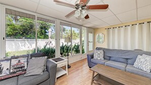 Un salon doté d’un ventilateur de plafond, d’un canapé gris, d’une table basse en bois et d’une grande fenêtre donnant sur un paysage verdoyant.