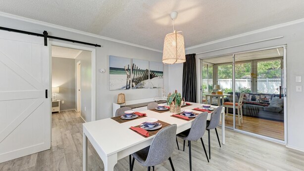 Un coin repas avec une table blanche dressée pour quatre personnes, un luminaire suspendu et une vue sur l’extérieur grâce à de grandes portes-fenêtres coulissantes.