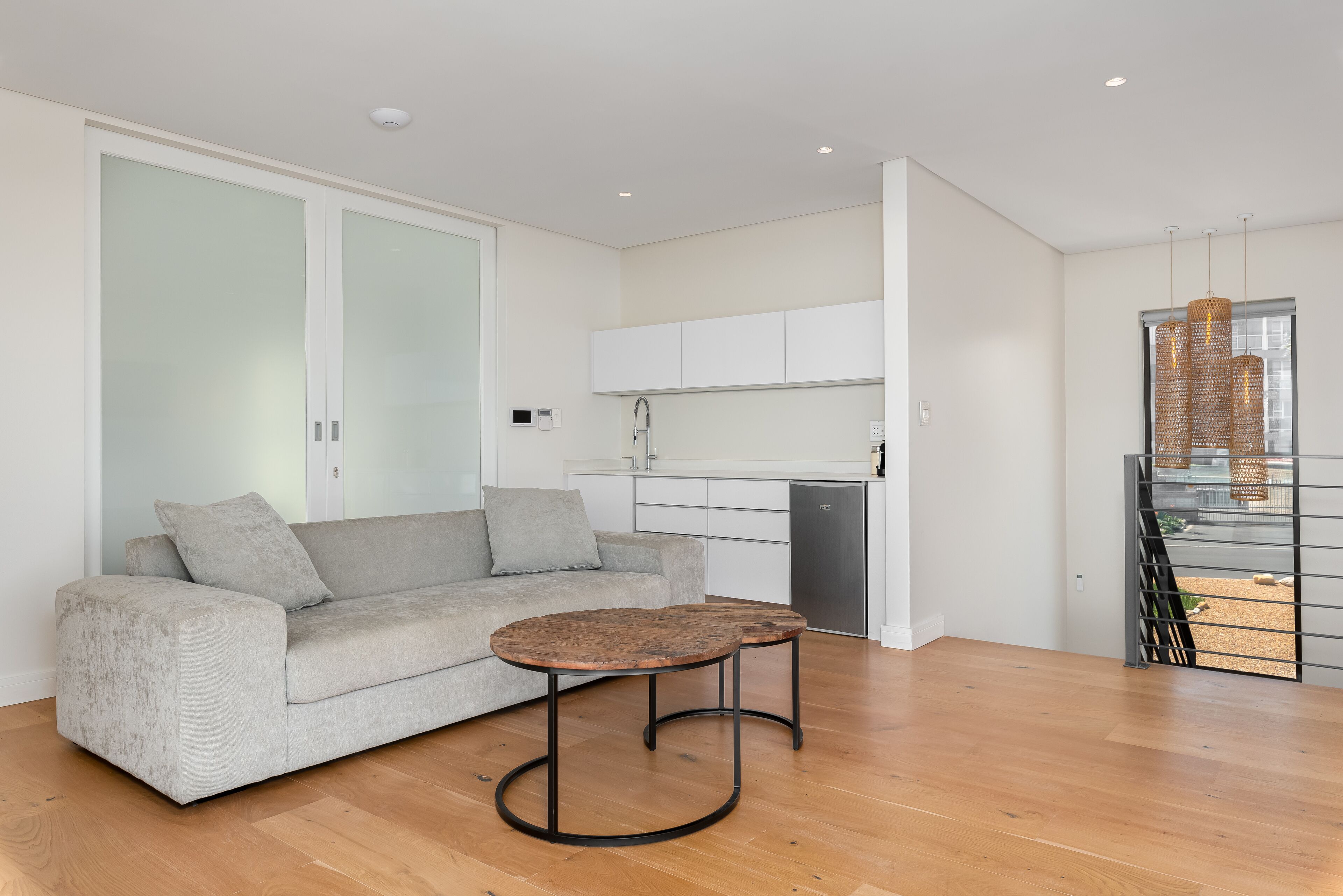 A modern apartment with a grey sofa, two round tables, a kitchenette, and a balcony with a view.