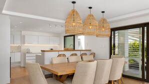 Et moderne spiseområde med et træbord og beige polstrede stole, et køkken i baggrunden og en stor skydedør i glas til højre.