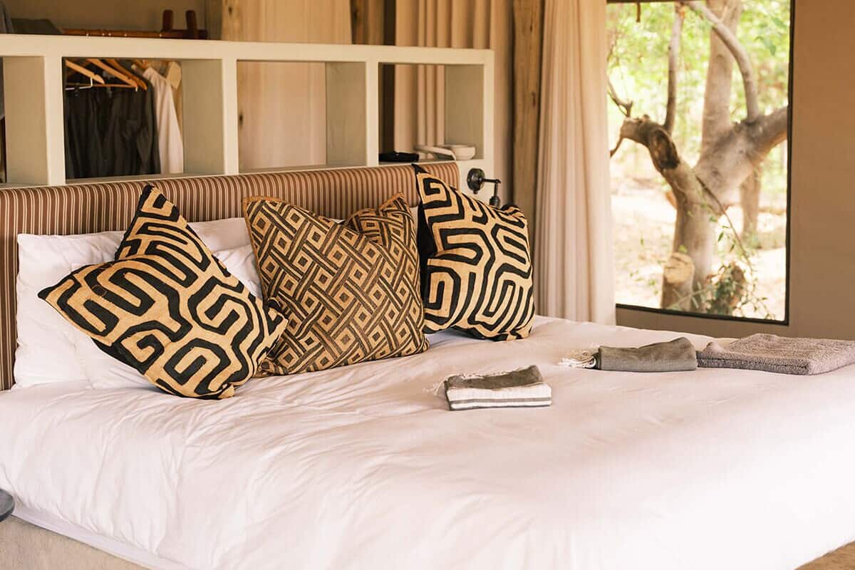 A bedroom with a bed, pillows, a bedside table, a window, and a view of trees.