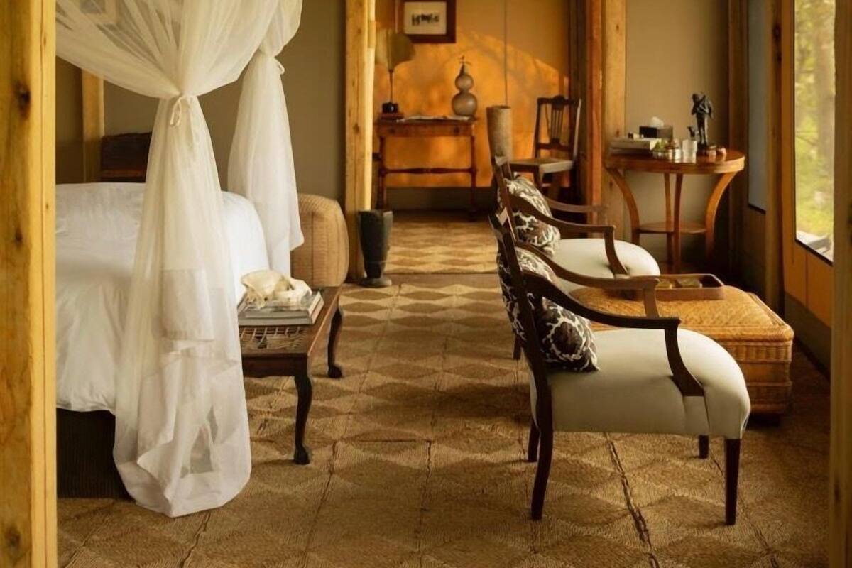 A bedroom with a canopy bed, a sitting area with chairs, a small table, and a view through a window.