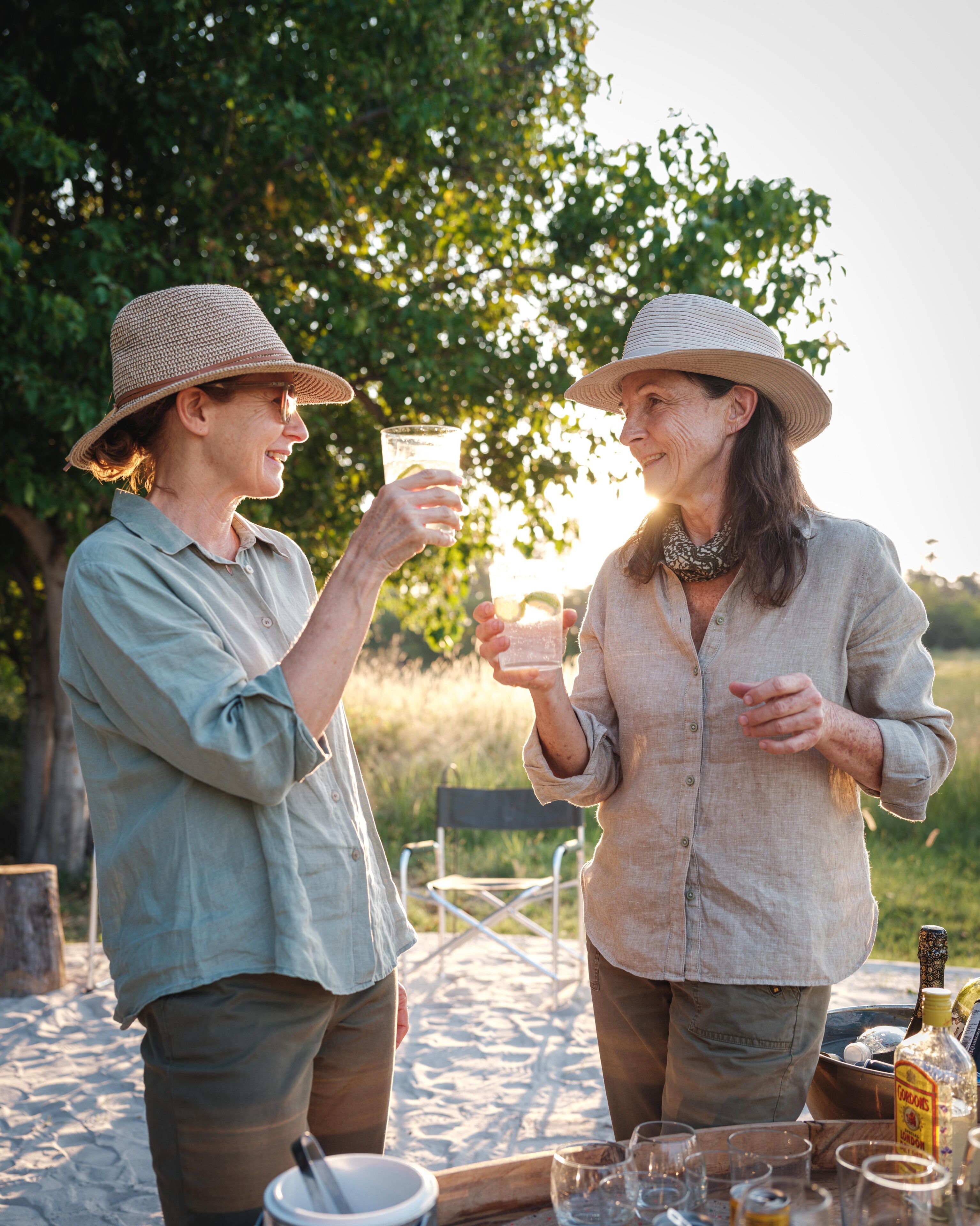 Two people toasting with drinks outdoors, surrounded by a picnic setup with a bottle and glasses on a table.
