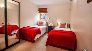 Une chambre d’hôtel avec deux lits, chacun recouvert d’une couverture rouge, une table de chevet avec une lampe et une fenêtre avec un store marron.