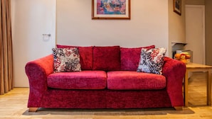 Un canapé en velours rouge agrémenté de coussins à motifs, une œuvre d’art florale encadrée accrochée au mur et un panier de fruits posé sur une table en bois.