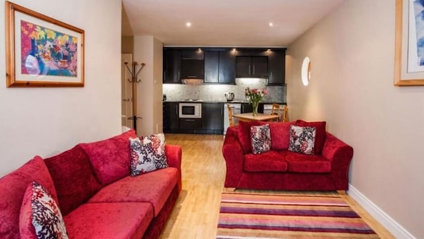 Un espace de vie compact avec deux canapés rouges, un tapis à rayures et une cuisine en arrière-plan.
