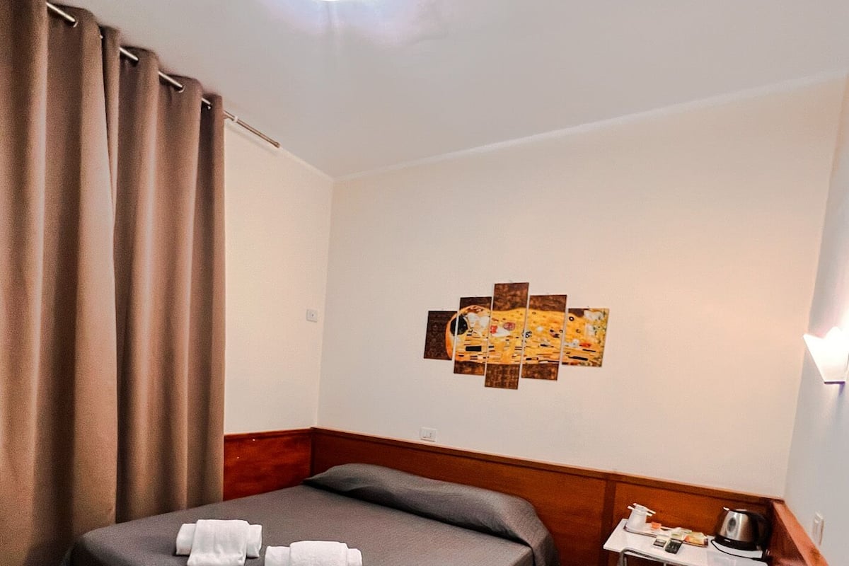 A hotel room with a bed, a small table with a kettle, and a wall-mounted artwork.