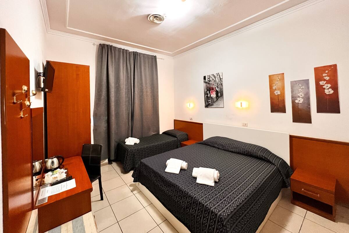 A hotel room with two beds, a desk with a kettle, a wardrobe, and a large window with curtains.
