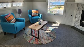 A room with two blue armchairs, a wooden coffee table, a patterned rug, and a view of a body of water through a large window. - Spacious lakeview home within minutes of beaches, state parks and shopping. (SPRING LAKE)