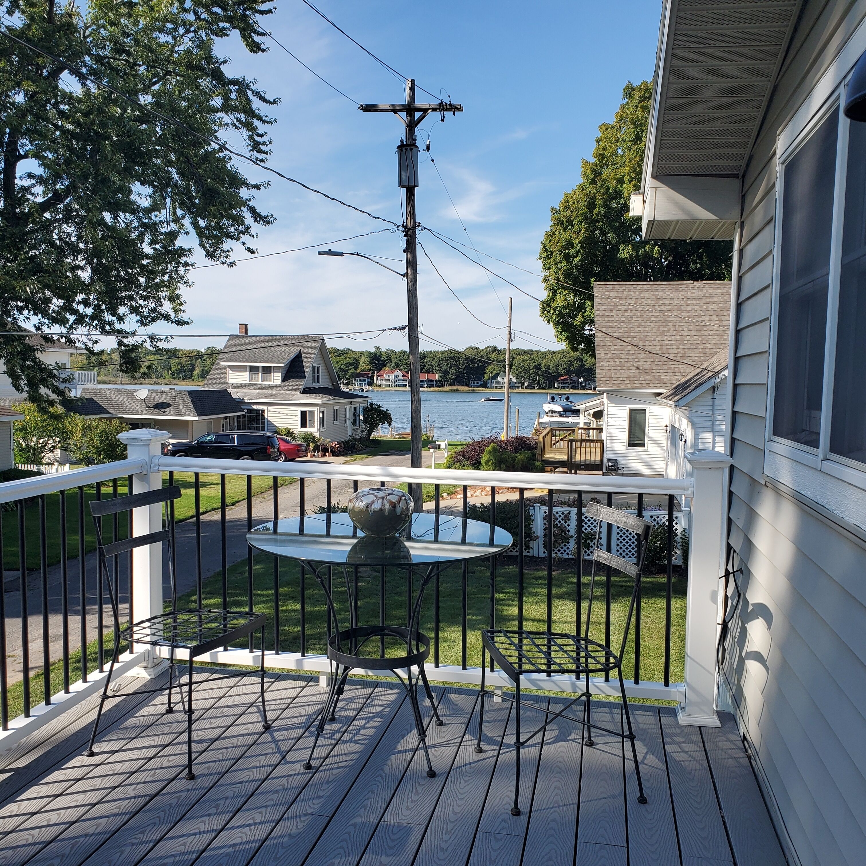 A wooden deck with a table and chairs, overlooking a lake with houses in the background.