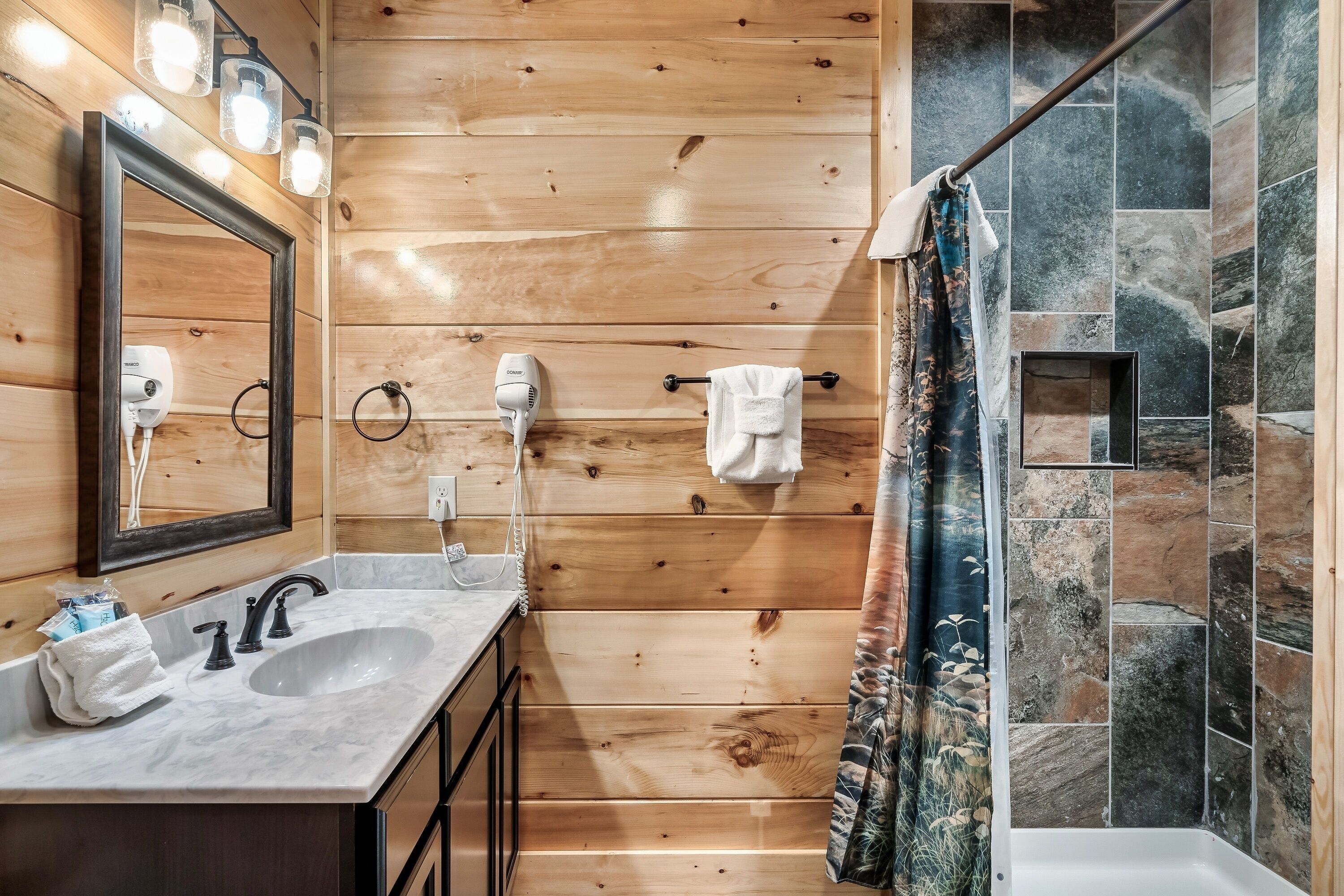 A wooden-paneled bathroom with a marble sink, two hair dryers, a mirror, and a shower with a stone wall.