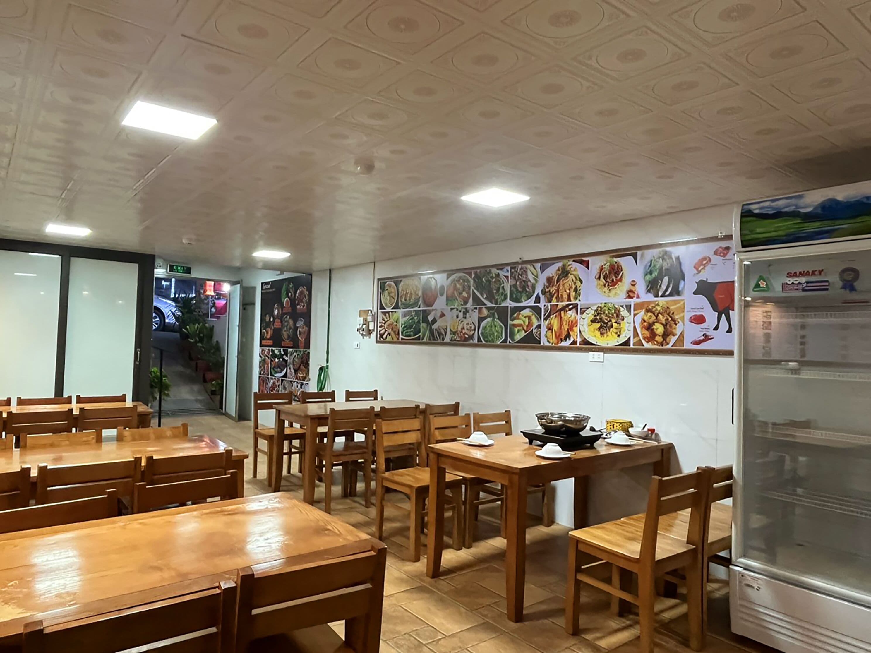 A restaurant interior with wooden tables and chairs, a wall with food images, and a refrigerator.