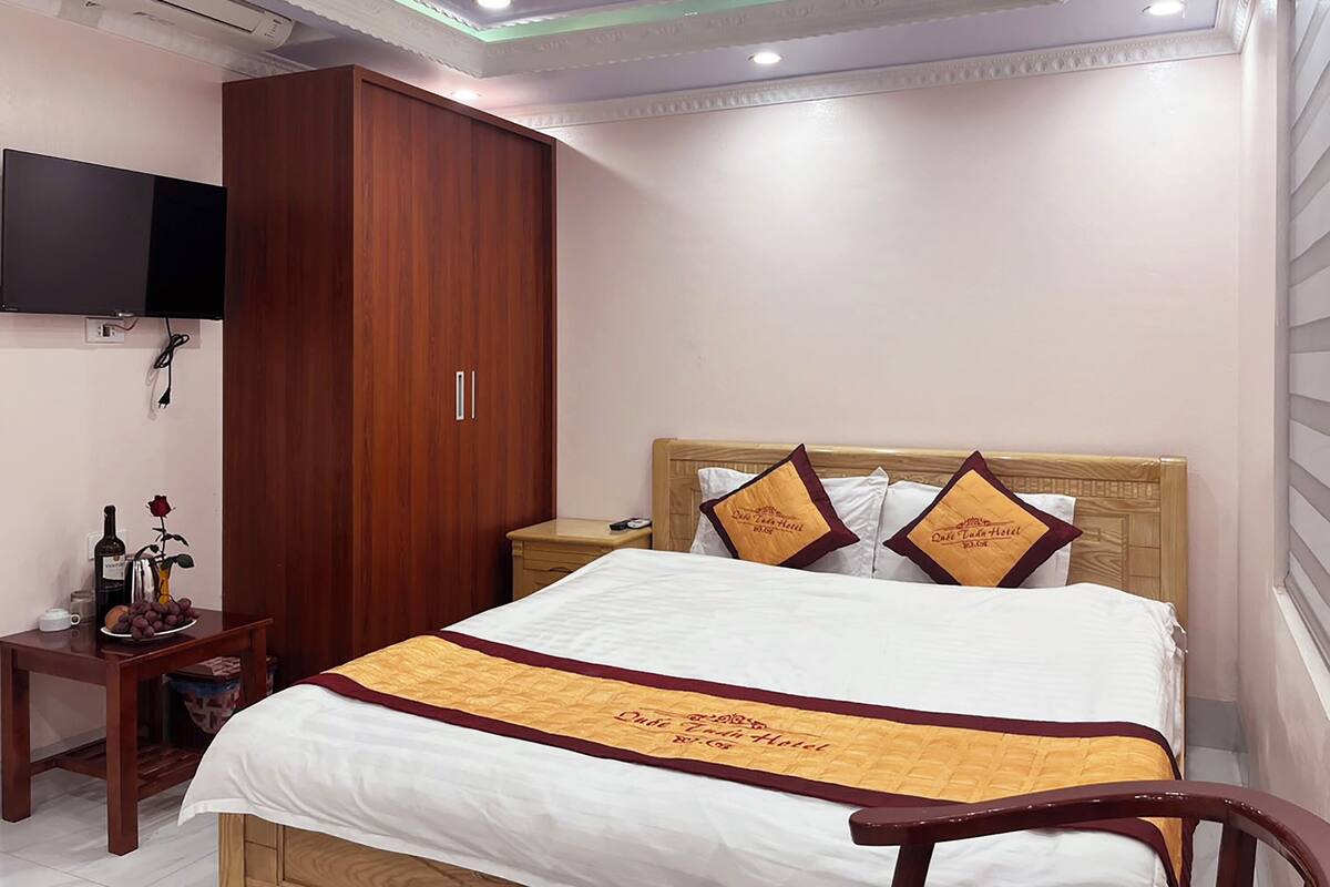 Quarto de hotel com cama, guarda-roupa de madeira, mesa de cabeceira, televisão na parede e teto com iluminação embutida.