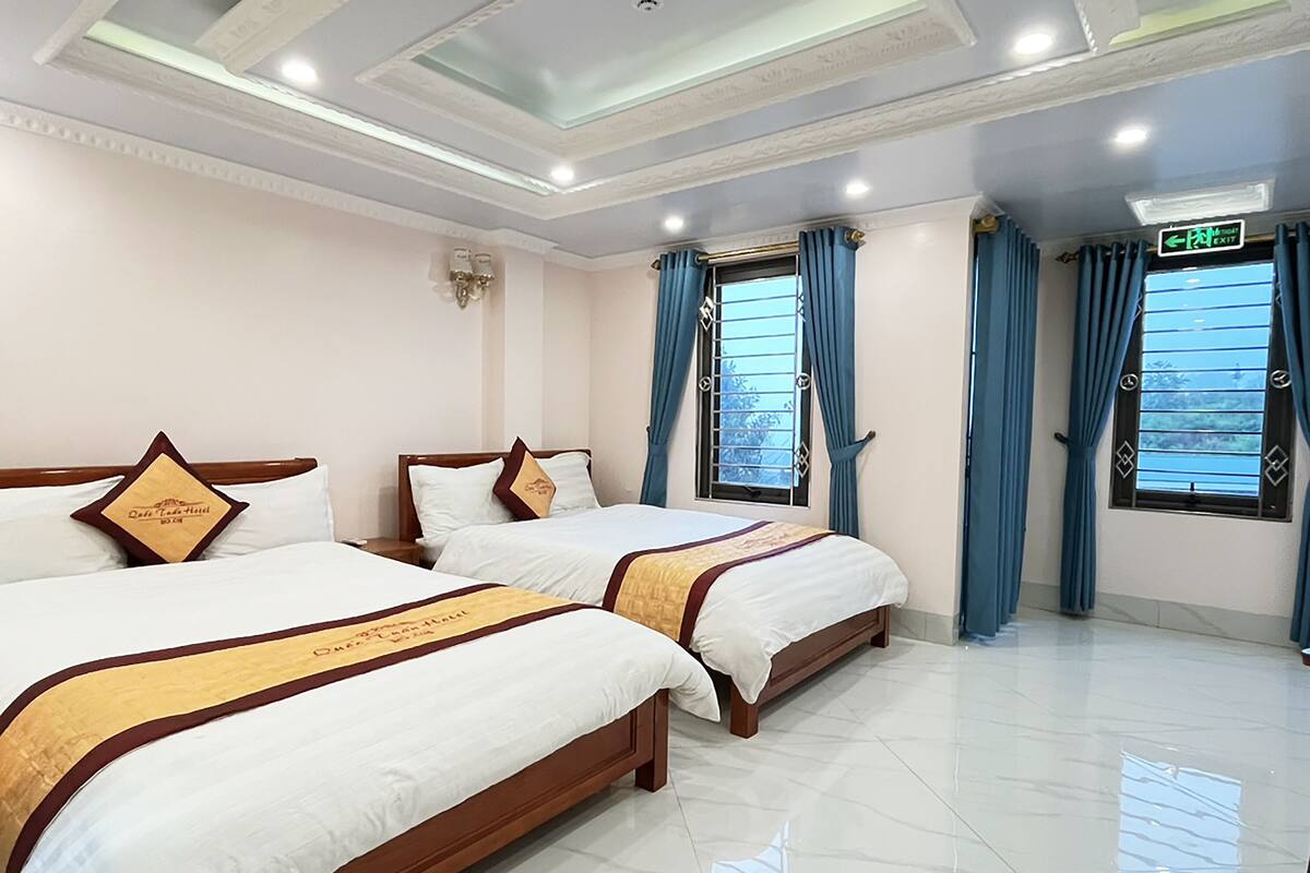 Quarto de hotel com duas camas, teto com iluminação embutida e janela com cortinas azuis.