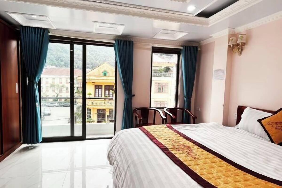 Quarto de hotel com cama, vista para edifícios e varanda com cadeiras.