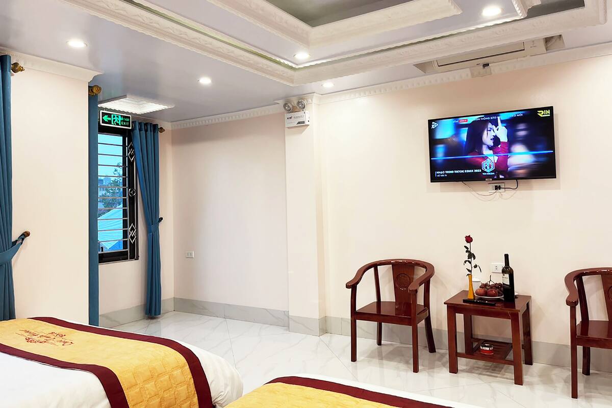 Quarto de hotel com cama, televisão fixada na parede, uma mesinha com garrafa e fruta, e uma cadeira.