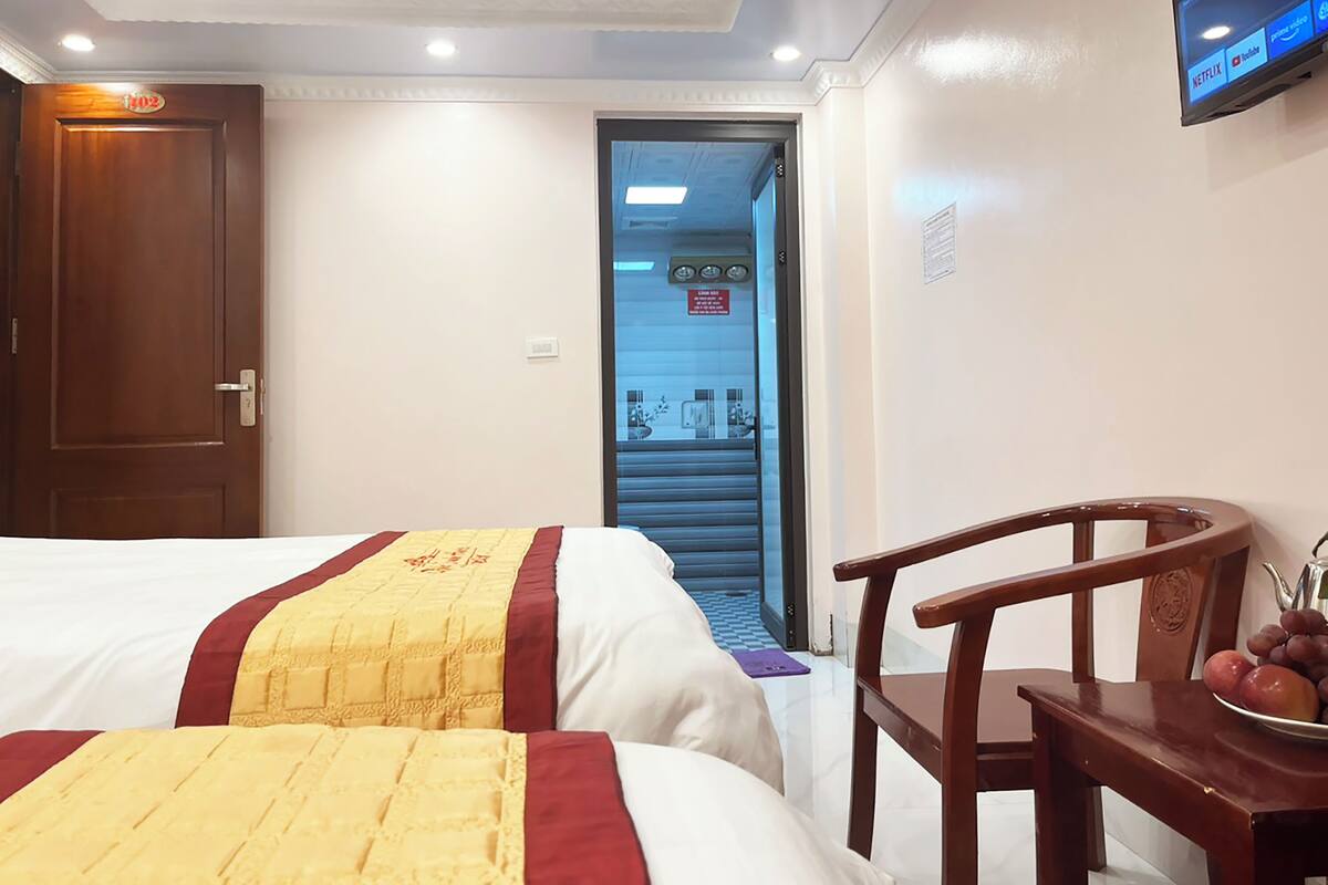 Quarto de hotel com duas camas, uma cadeira de madeira, uma mesa com frutas e uma televisão fixada na parede.