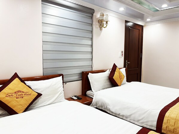 A hotel room with two beds, each with white bedding and a decorative pillow. There is a window with blinds, a wall-mounted light fixture, and a door. - Quoc Tuan Sapa Hotel (Sa Pa)