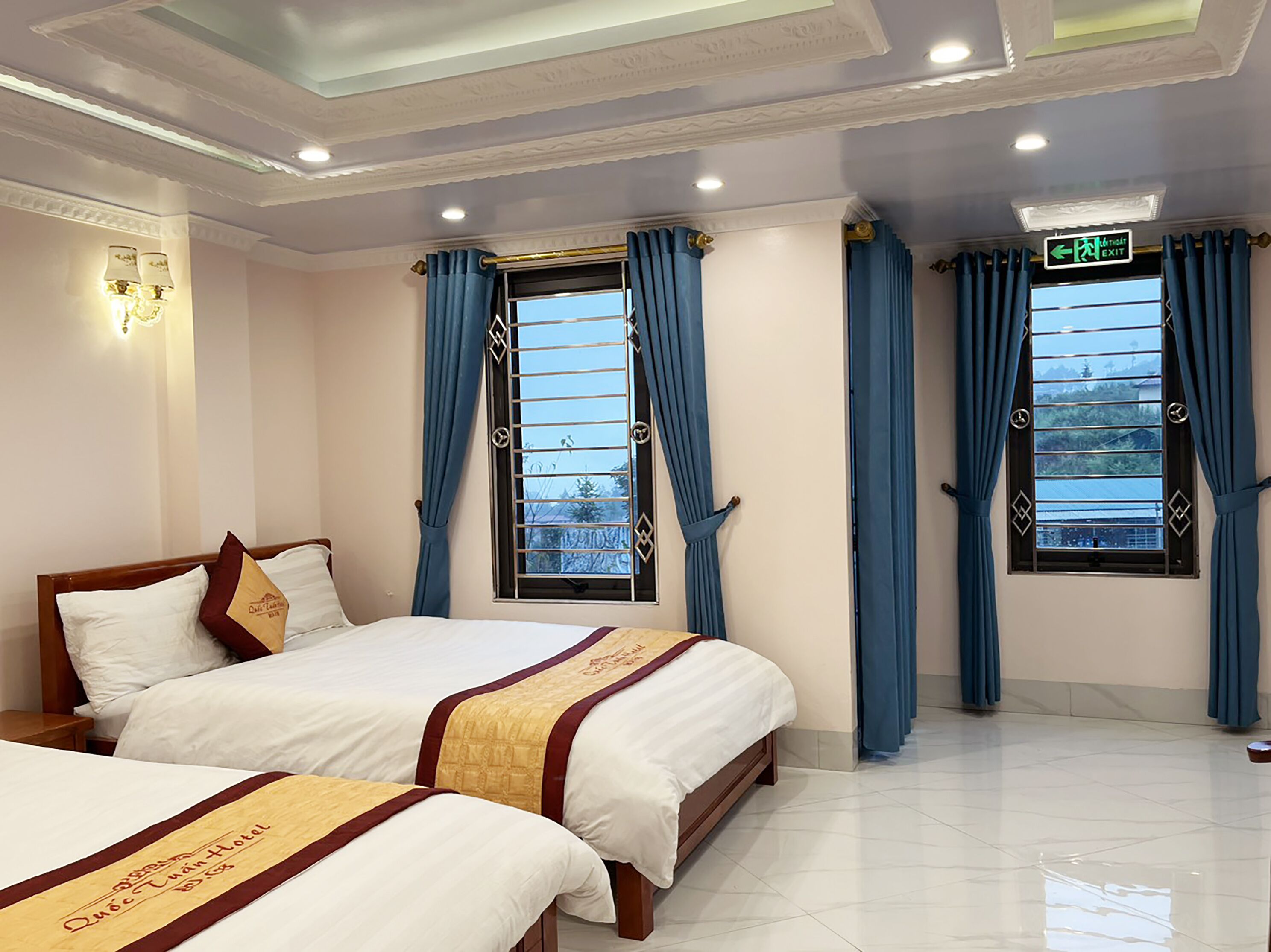 A hotel room with two beds, a ceiling with decorative molding, and windows with blue curtains.