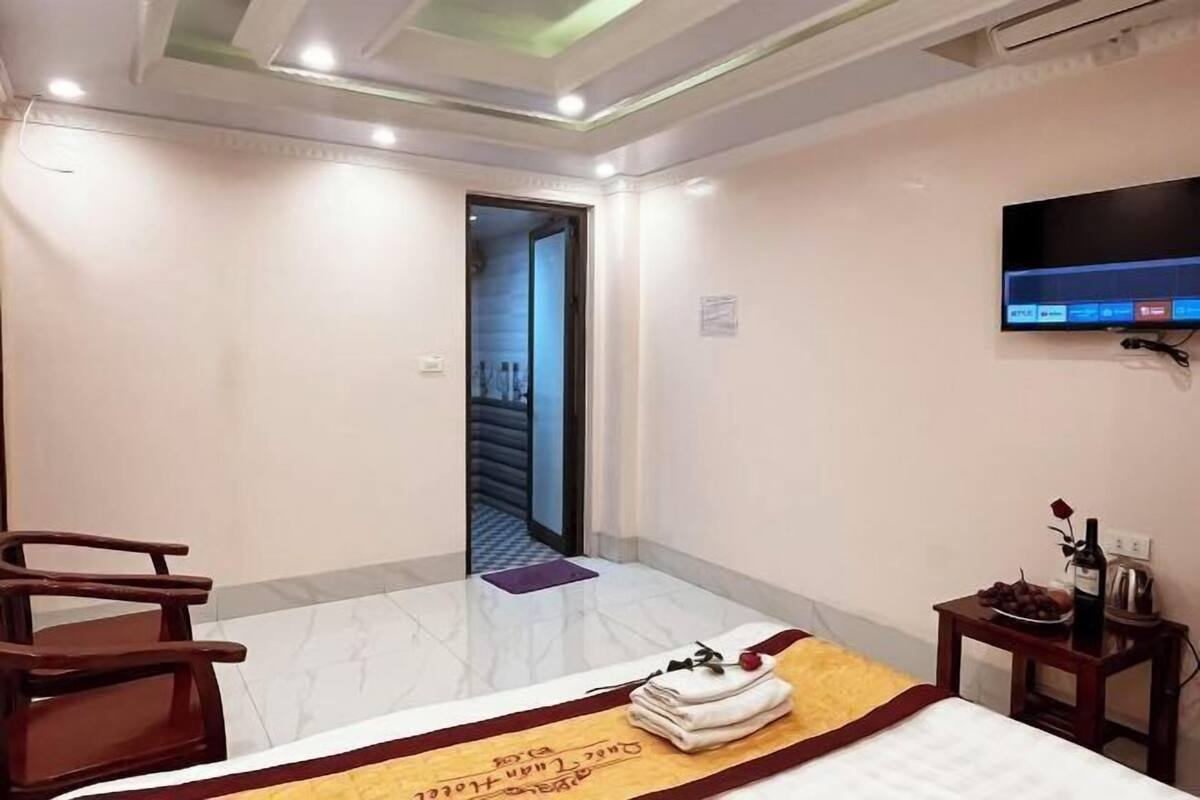 Quarto de hotel com cama, televisão, uma mesinha com um frasco e uma rosa, e uma cadeira.