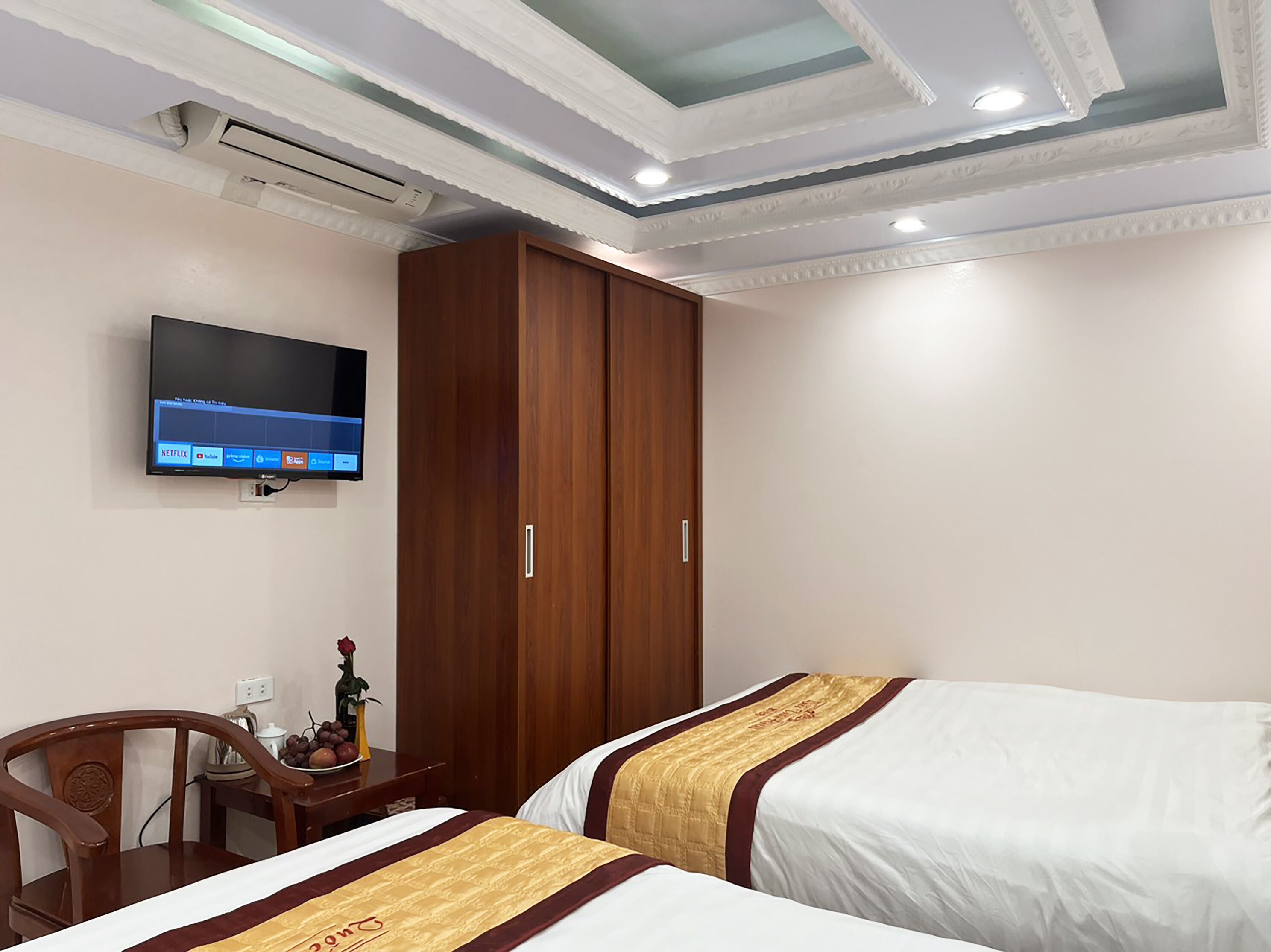 A hotel room with two beds, a wooden wardrobe, a TV mounted on the wall, and a small table with a vase of flowers.