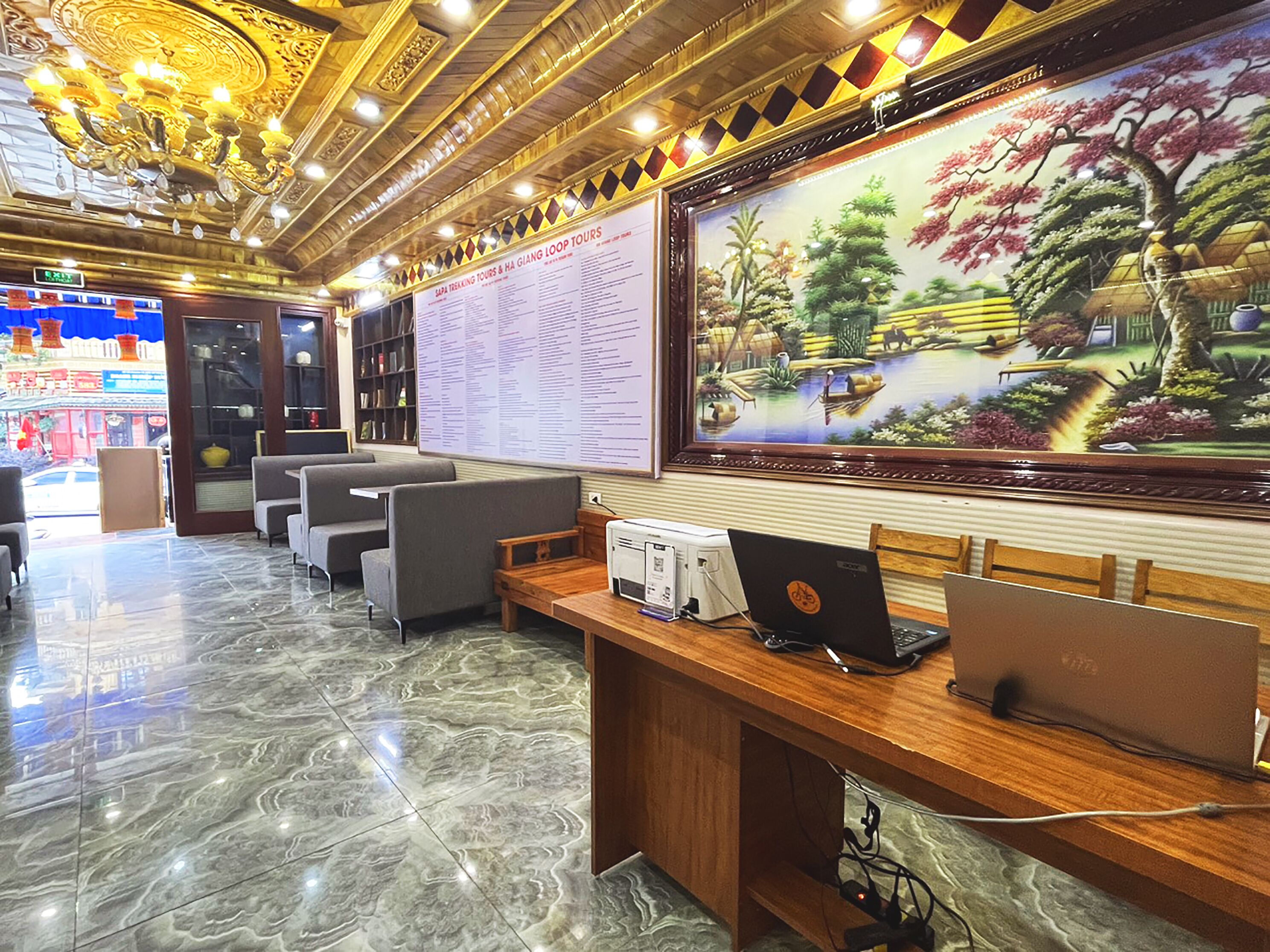A lobby with a wooden reception desk, two laptops, a large painting of a garden, and a wall with a list of tours.